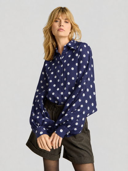 Eine Frau mit blonden Haaren trägt die Bluse „Antonia Dots Navy Ecru“ – eine übergroße, marineblaue Bluse mit langen Ärmeln und Polka-Dots aus weichem Baumwoll-Voile. Kombiniert mit dunklen Shorts steht sie vor einem schlichten, hellen Hintergrund und präsentiert dieses mühelos schicke Kleidungsstück, das stolz in Italien hergestellt wurde.
