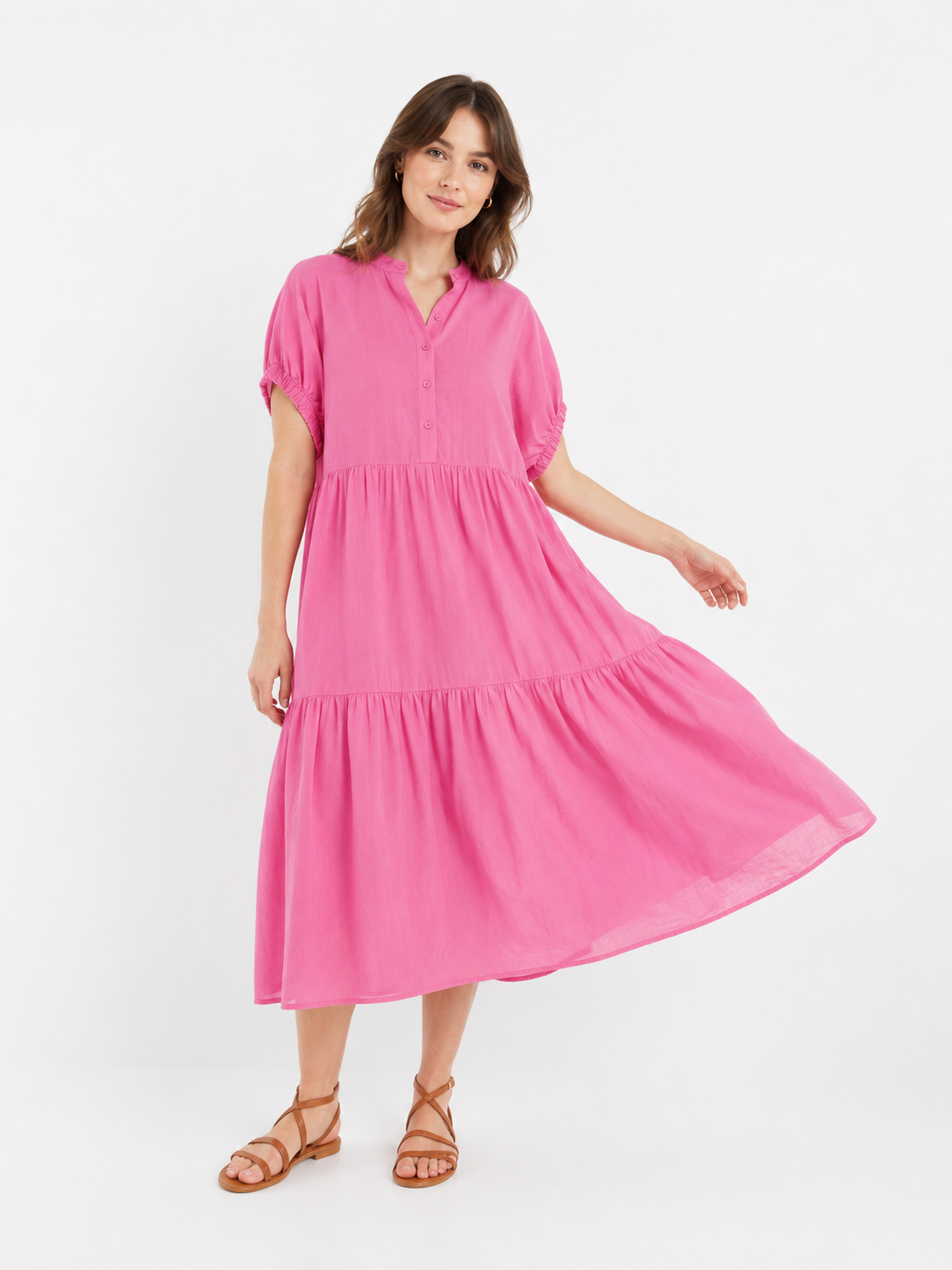 Eine Frau, die das „Cowboy Boho Pink“-Kleid trägt – ein leuchtend pinkfarbenes, kurzärmeliges, gestuftes Maxikleid aus weichem Baumwollvoile mit Knopfdetails am Oberteil –, steht vor einem schlichten weißen Hintergrund und hebt eine Seite des Rocks leicht an. Zu diesem „Cowboy Boho“-Kleid trägt sie braune Riemchensandalen.