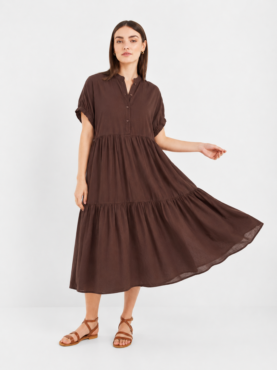 Eine Frau, die das „Dress Cowboy Boho Brown“ trägt – ein braunes, kurzärmeliges, geknöpftes Midi-Kleid mit Stufenschnitt –, posiert in Kombination mit braunen Sandalen vor einem weißen Hintergrund und hält dabei eine Seite des Kleides hoch.