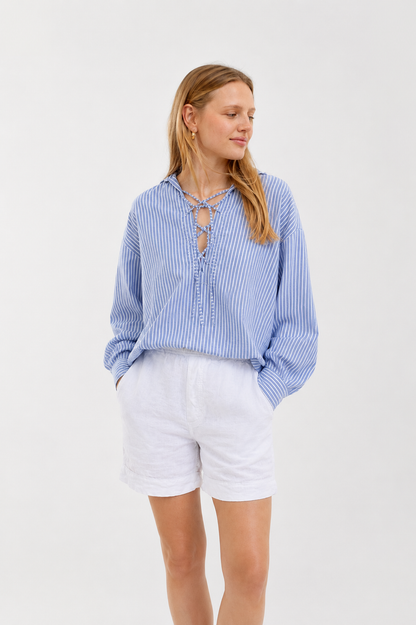 Vor einem schlichten Hintergrund steht eine Frau mit langen Haaren. Sie trägt die Bluse „Fisherman Light Blue Stripes“ – eine blau-weiß gestreifte Bluse mit Schnürung am Ausschnitt – dazu weiße Shorts. Die Bluse wurde in Italien aus 100 % Baumwolle hergestellt; die Frau blickt mit ausdruckslosem Gesicht zur Seite.