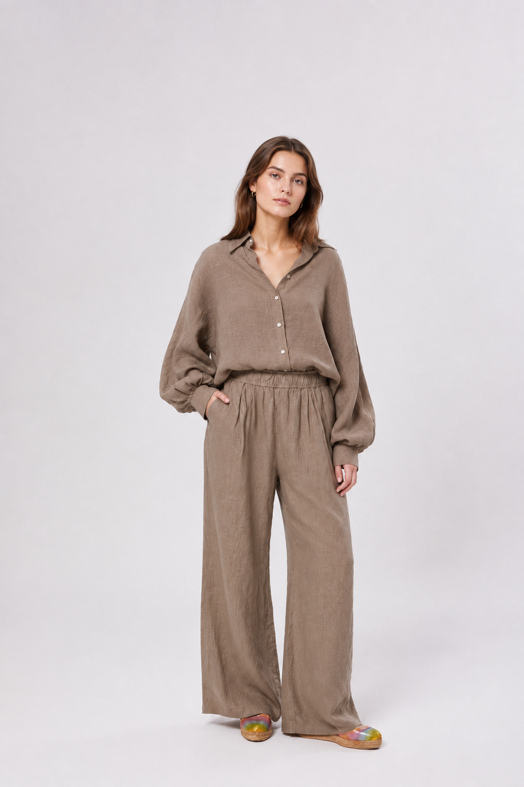 Eine Frau steht vor einem hellgrauen Hintergrund und trägt ein locker sitzendes Oberteil „Antonia Linen Soft Brown“ sowie eine dazu passende Hose mit weitem Bein; ihre linke Hand steckt in der Tasche. Sie hat langes braunes Haar und trägt bunte Schuhe.