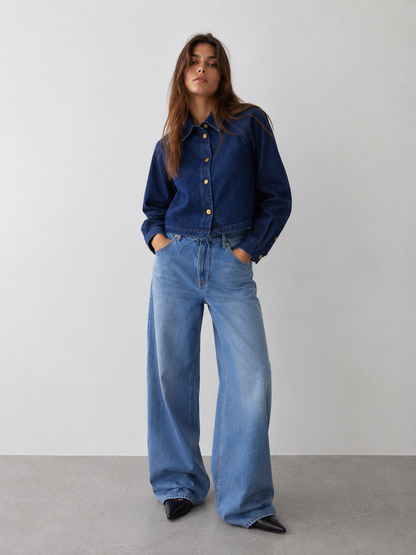 Vor einem schlichten Hintergrund steht eine Frau, die die Jacke „Hanna Denim“ in kastenförmiger Passform und eine locker sitzende hellblaue Jeans sowie schwarze spitze Schuhe trägt und selbstbewusst in die Kamera blickt.