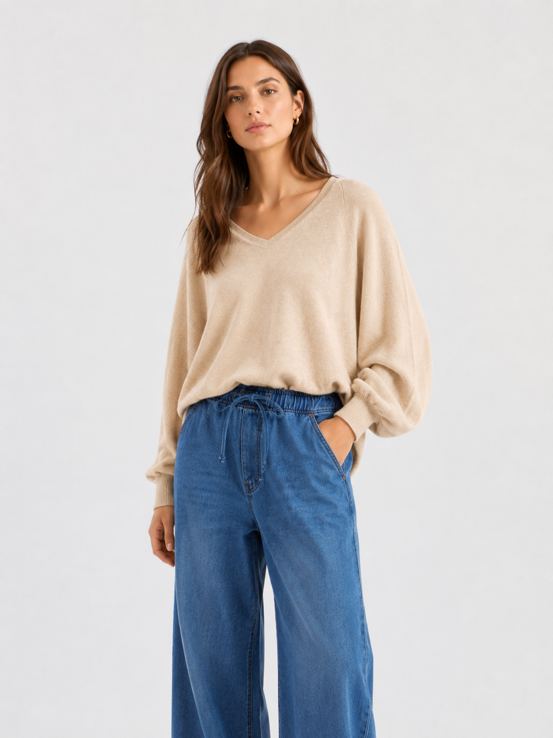 Eine Person steht vor einem schlichten Hintergrund und trägt einen Pullover „Antonia V-Neck Puder“ aus weicher Merinowolle und Kaschmir mit Ballonärmeln, dazu eine hoch taillierte Bluejeans mit weitem Bein.