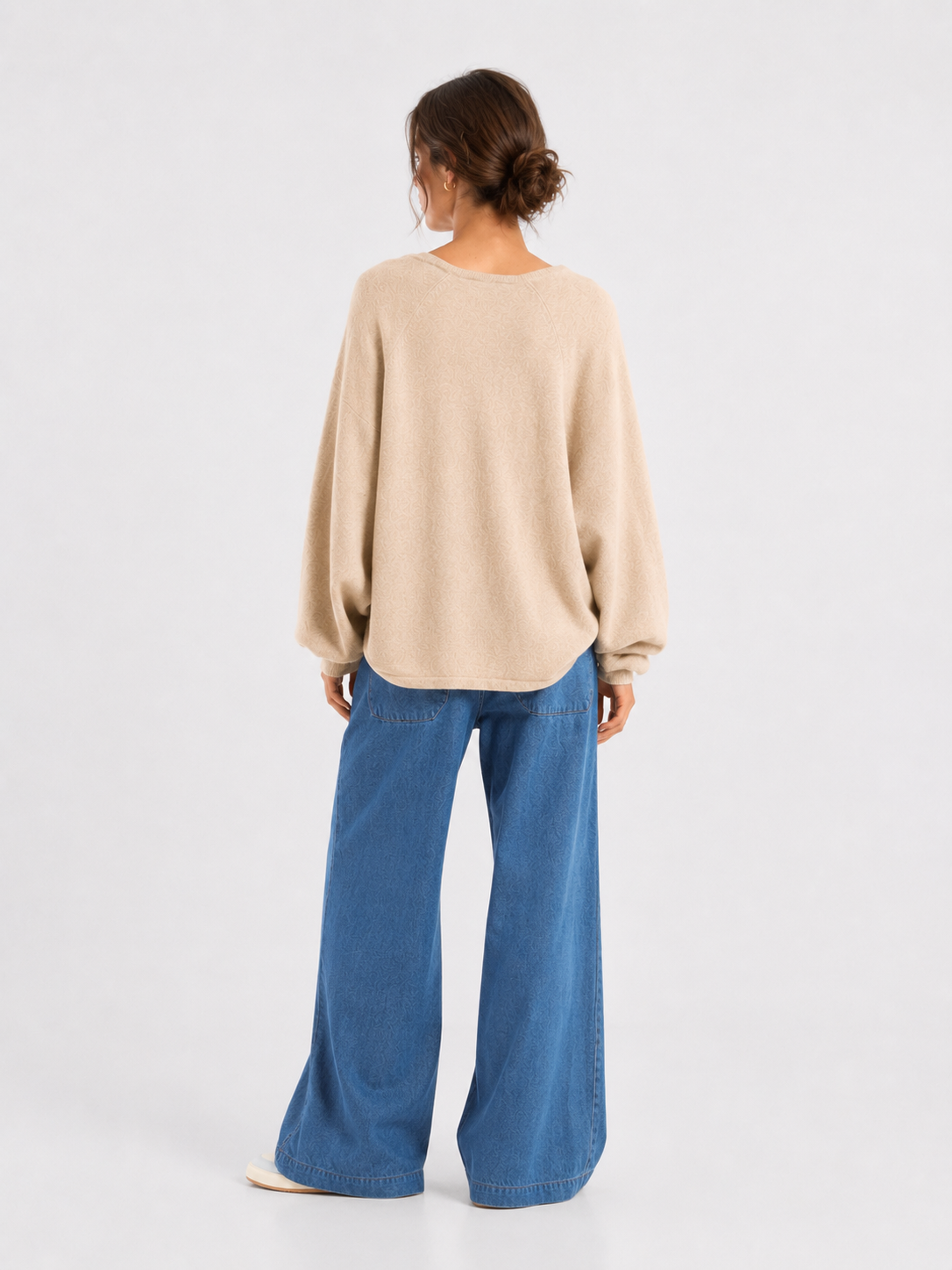 Eine Frau steht mit dem Rücken zur Kamera und trägt einen lockeren beigen Pullover, eine Hose „Anna“ aus Denim mit weitem Bein und elastischem Bund sowie weiße Schuhe. Ihr Haar ist zu einem tief sitzenden Dutt gesteckt, und der Hintergrund ist schlicht und hell.