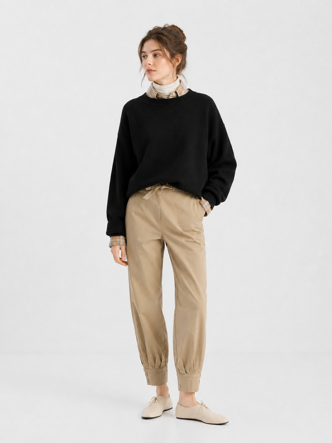 Vor einem schlichten Hintergrund steht eine Frau, die den Pullover „Giulia Schwarz“ über einem karierten Hemd, einer hellbraunen Hose mit Umschlag und beigen Schuhen trägt. Sie blickt zur Seite und hat eine Hand in der Tasche.