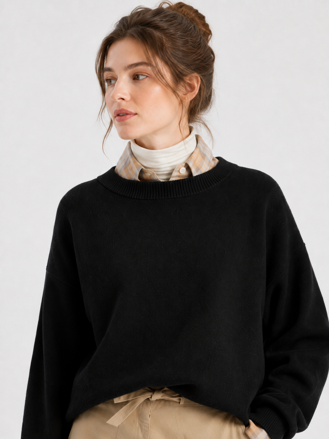 Eine Frau mit braunen Haaren, die zu einem Dutt zusammengebunden sind, trägt den Pullover „Giulia Schwarz“ über einem karierten Hemd und einem weißen Rollkragenpullover und blickt vor einem einfarbigen Hintergrund zur Seite.
