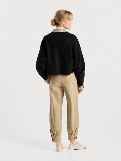 Eine Frau steht mit dem Rücken zur Kamera und trägt den Pullover „Giulia Schwarz“ über einem karierten Hemd, dazu beige Hosen mit geknöpften Bündchen und helle Schuhe vor einem schlichten weißen Hintergrund.