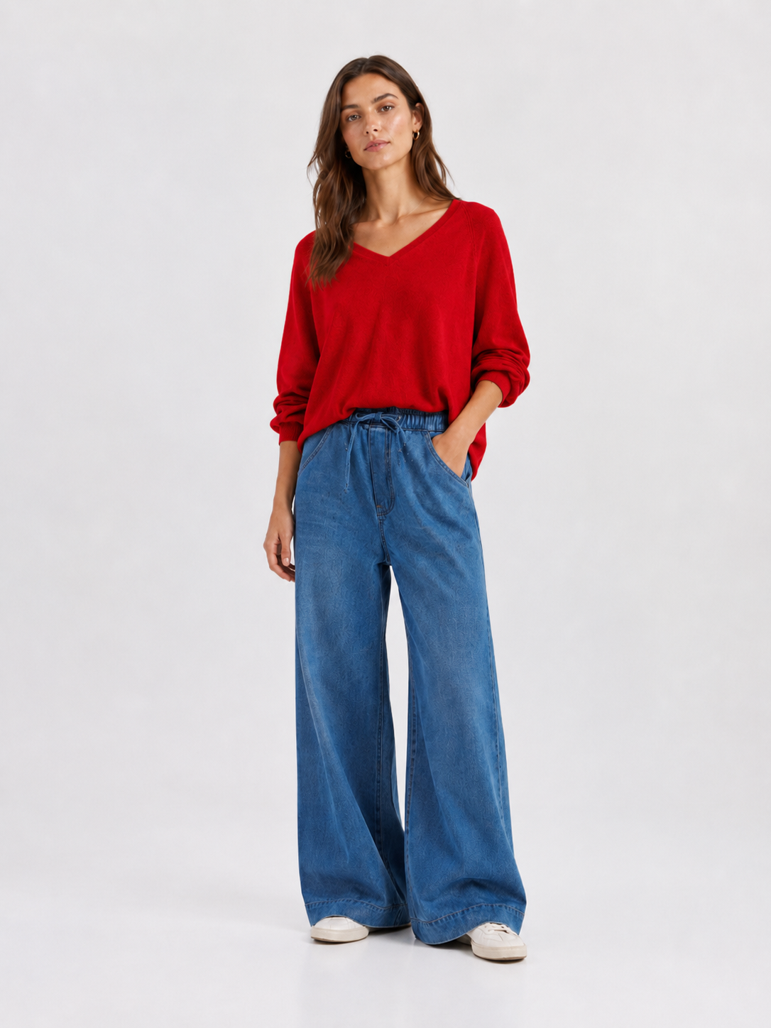 Eine Frau steht mit dem Blick nach vorne und trägt den Pullover „Antonia V-Neck Rot“ aus weicher Merinowolle und Kaschmir, dazu eine blaue Jeans mit weitem Bein und weiße Schuhe. Der Hintergrund ist einfarbig und hell.