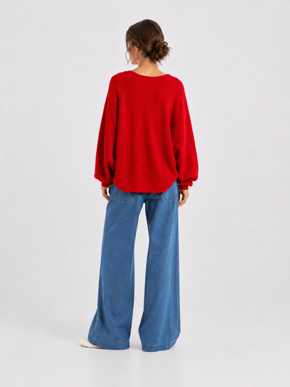 Eine Person steht mit dem Rücken zur Kamera und trägt einen locker sitzenden Pullover „Antonia V-Neck Rot“ aus weicher Merinowolle und Kaschmir, dazu eine blaue Jeans mit weitem Bein. Der Hintergrund ist einfarbig und hell.