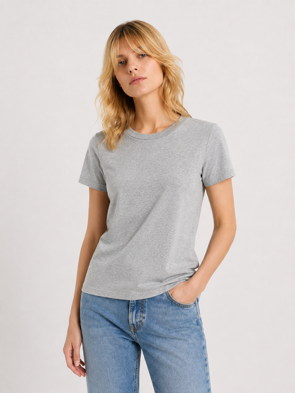 Eine Person mit blonden Haaren, die ein graues T-Shirt mit Rundhalsausschnitt und eine blaue Jeans trägt, steht vor einem schlichten hellen Hintergrund und hat eine Hand in der Hosentasche.