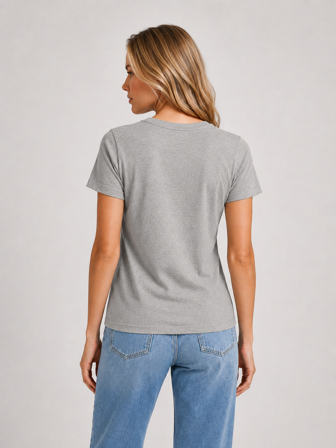 Eine Frau mit blonden Haaren, die ein graues, kurzärmeliges T-Shirt mit Rundhalsausschnitt und eine blaue Jeans trägt, steht mit dem Rücken zur Kamera vor einem schlichten, hellen Hintergrund. Dieses stilvolle Shirt wird in Europa aus hochwertiger Baumwolle gefertigt und überzeugt durch seine langlebige Qualität.