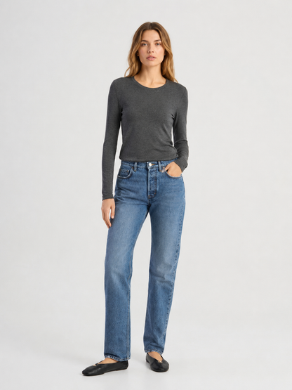 Vor einem schlichten Hintergrund steht eine Frau, die das langärmelige Kaschmir-Shirt „Cashmere Long Sleeve Rib“ in Dunkelgrau trägt, kombiniert mit einer blauen Jeans und schwarzen Ballerinas. Eine Hand steckt in ihrer Hosentasche, während sie mit ausdruckslosem Gesicht nach vorne blickt. Das in Portugal gefertigte Shirt besticht durch seinen lässigen Stil und seine raffinierte europäische Handwerkskunst.
