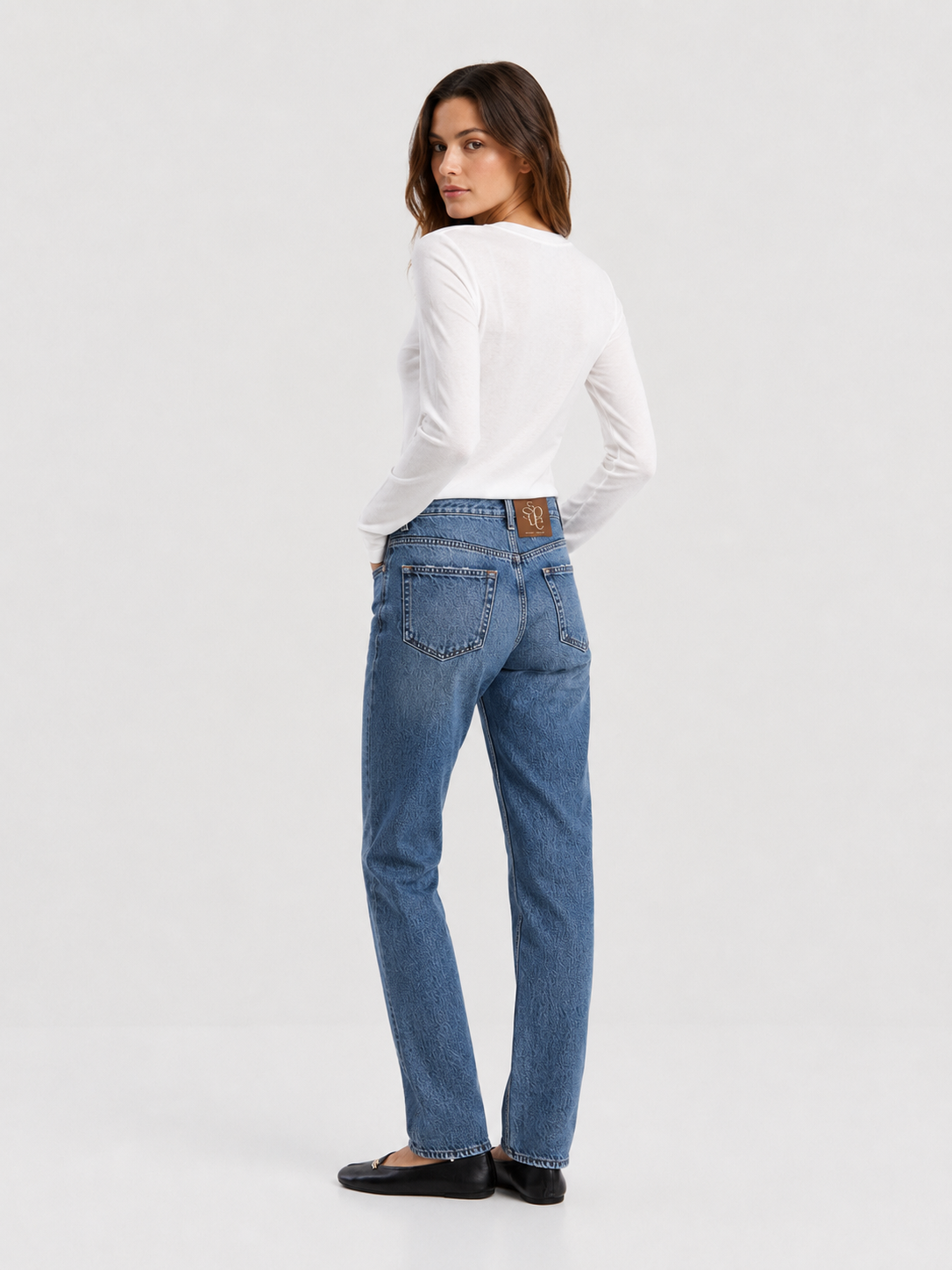 Eine Frau steht mit dem Rücken zur Kamera und trägt ein weißes, langärmeliges Cashmere-Oberteil mit V-Ausschnitt und Rippbündchen sowie eine blaue Jeans und schwarze Ballerinas. Sie hat die linke Hand in der Tasche und blickt über ihre linke Schulter. Der Hintergrund ist einfarbig und hell.