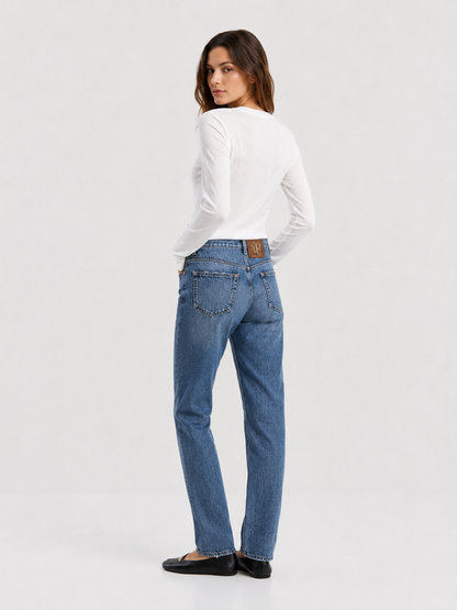 Eine Frau steht mit dem Rücken zur Kamera und trägt ein weißes, langärmeliges Cashmere-Oberteil mit V-Ausschnitt und Rippbündchen sowie eine blaue Jeans und schwarze Ballerinas. Sie hat die linke Hand in der Tasche und blickt über ihre linke Schulter. Der Hintergrund ist einfarbig und hell.
