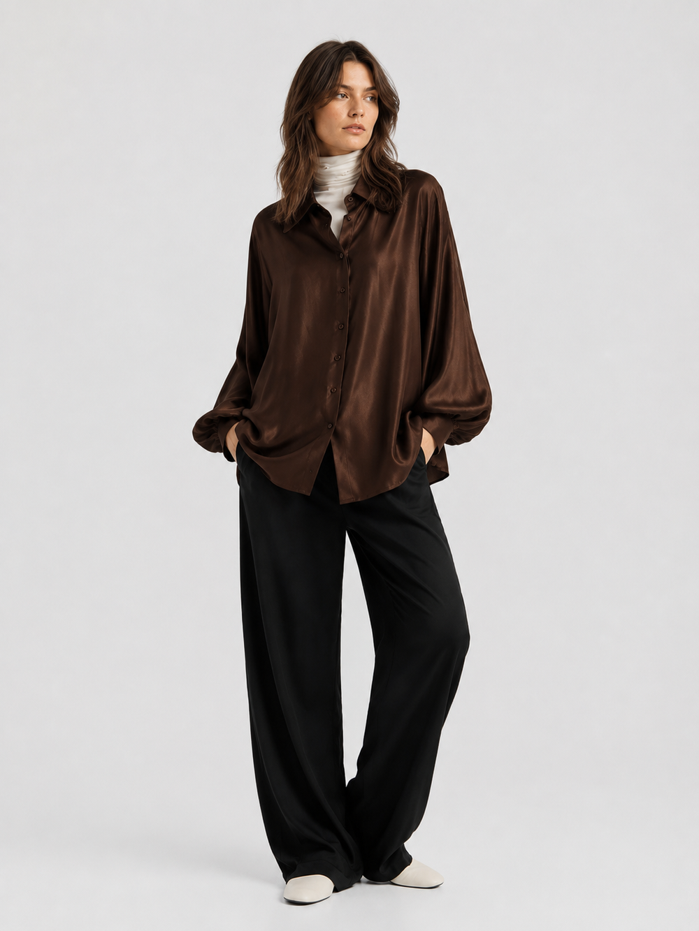 Eine Frau steht mit den Händen in den Taschen vor einem schlichten, hellen Hintergrund. Sie trägt das braune Satinhemd mit Knöpfen von „Espresso Hour“ über einem weißen Rollkragenpullover, dazu eine weite schwarze Hose und weiße Schuhe.