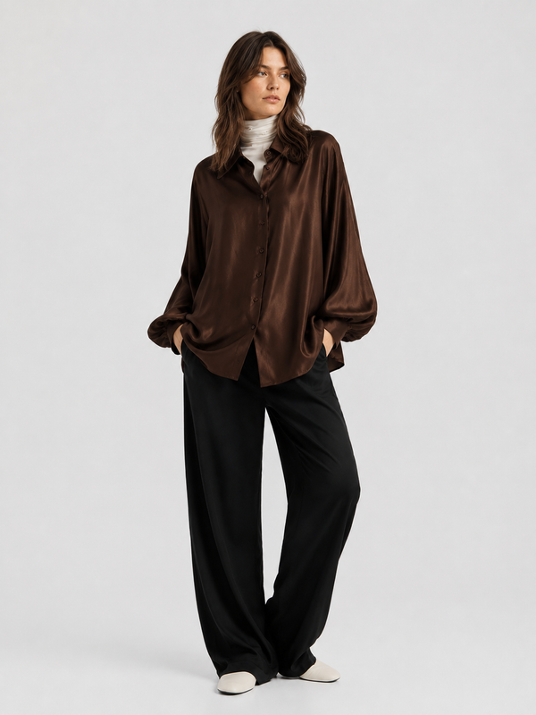 Eine Frau steht mit den Händen in den Taschen vor einem schlichten, hellen Hintergrund. Sie trägt das braune Satinhemd mit Knöpfen von „Espresso Hour“ über einem weißen Rollkragenpullover, dazu eine weite schwarze Hose und weiße Schuhe.