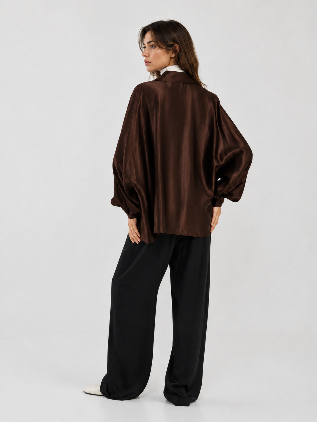 Eine Frau steht mit dem Rücken zur Kamera und trägt die „Bluse Antonia Shiny Brown One Size“ – eine locker sitzende braune Bluse –, kombiniert mit einer weiten schwarzen Hose und weißen Schuhen vor einem schlichten hellen Hintergrund. Dieser stilvolle Look wurde in Portugal hergestellt.