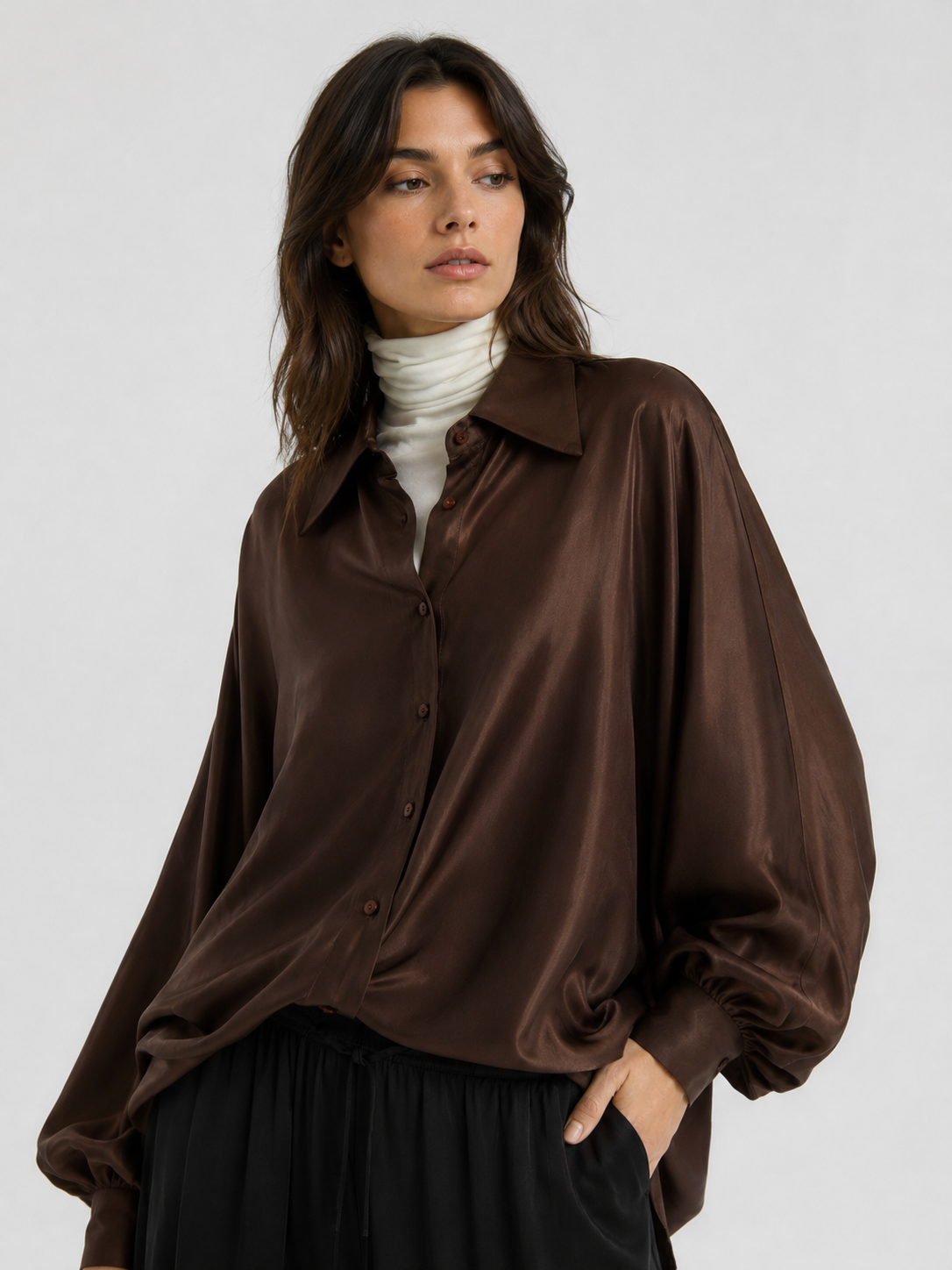 Eine Person trägt die Bluse „Antonia Shiny Brown One Size“ – ein glänzendes, oversize-Schnitt-Hemd mit Knopfleiste, das in Portugal gefertigt wurde – über einem weißen Rollkragenpullover, steht mit einer Hand in der Tasche da und blickt vor einem schlichten, hellen Hintergrund zur Seite. Dieses vielseitige Kleidungsstück verleiht jedem Outfit mühelosen Stil und Raffinesse.