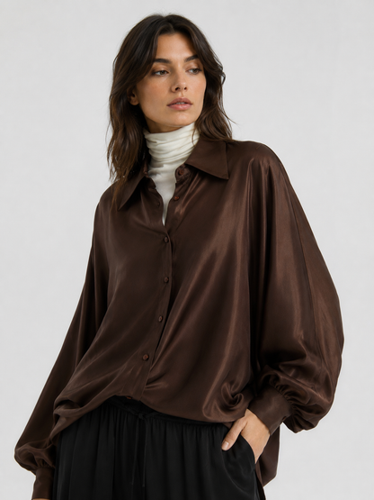 Eine Person trägt die Bluse „Antonia Shiny Brown One Size“ – ein glänzendes, oversize-Schnitt-Hemd mit Knopfleiste, das in Portugal gefertigt wurde – über einem weißen Rollkragenpullover, steht mit einer Hand in der Tasche da und blickt vor einem schlichten, hellen Hintergrund zur Seite. Dieses vielseitige Kleidungsstück verleiht jedem Outfit mühelosen Stil und Raffinesse.
