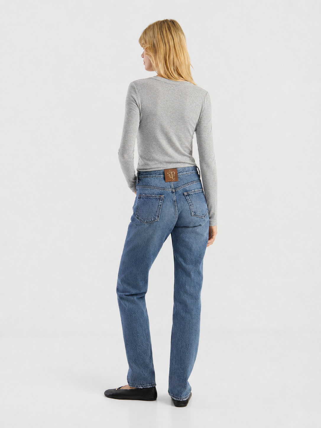 Eine Person mit blonden Haaren, die ein graues Langarmshirt, eine blaue Jeans und schwarze Schuhe trägt, steht mit dem Rücken zur Kamera vor einem schlichten weißen Hintergrund. Das Shirt wurde in Portugal hergestellt.
