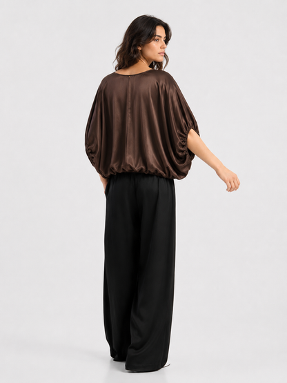Eine Frau steht in einem braunen, locker sitzenden Satin-Oberteil mit Fledermausärmeln und einer schwarzen Hose mit weitem Bein – das Oberteil ist ein Modell „Bluse Cowboy Shiny Braun“ aus LENZING ECOVERO-Viskose – und blickt mit leicht zur Seite gedrehtem Kopf von einem einfarbigen Hintergrund weg.
