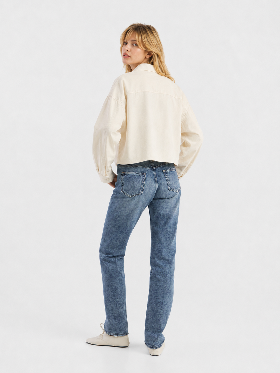 Eine Person mit blonden Haaren steht mit dem Rücken zur Kamera und trägt eine cremefarbene, verkürzte Flanellbluse, blaue Jeans und weiße Schuhe vor einem schlichten weißen Hintergrund.