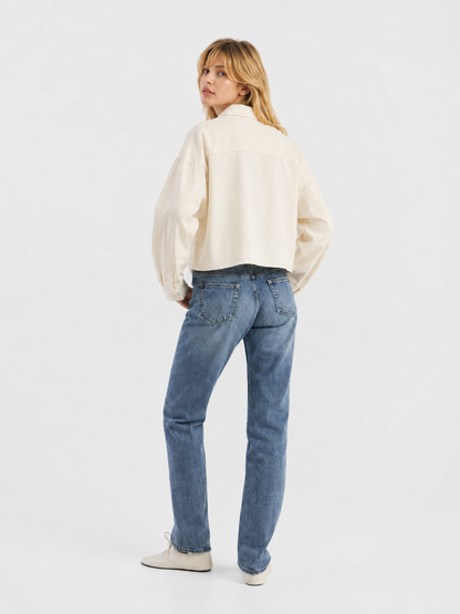 Eine Person mit blonden Haaren steht mit dem Rücken zur Kamera und trägt eine cremefarbene, verkürzte Flanellbluse, blaue Jeans und weiße Schuhe vor einem schlichten weißen Hintergrund.