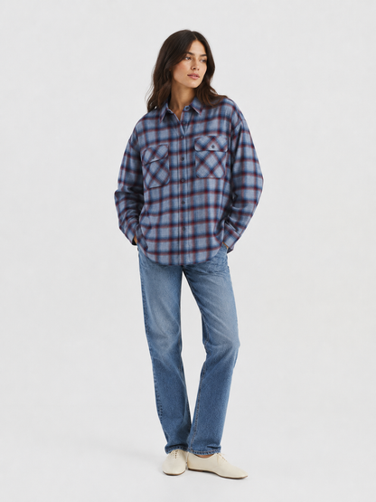 Vor einem schlichten, hellen Hintergrund steht eine Person mit den Händen in den Taschen und trägt das blau-rote Karomuster-Hemd von Ted Flanell sowie blaue Jeans und weiße Schuhe.