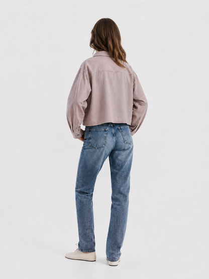 Vor einem schlichten weißen Hintergrund steht eine Person mit braunen Haaren mit dem Rücken zur Kamera. Sie trägt die Jacke „Flanell Cropped Taupe“, die wie ein Hemd geschnitten ist, eine blaue Jeans und beige Schuhe.