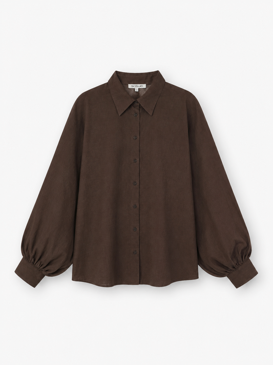 Die Bluse „Antonia Slim Espresso“ ist eine dunkelbraune, langärmelige Bluse mit Knopfleiste, spitzem Kragen und voluminösen Ballonärmeln, die an den Manschetten gerafft sind. Sie ist aus hochwertiger Baumwolle gefertigt und wird vor einem weißen Hintergrund präsentiert. Diese elegante Bluse wird in Portugal in meisterhafter Handarbeit hergestellt.