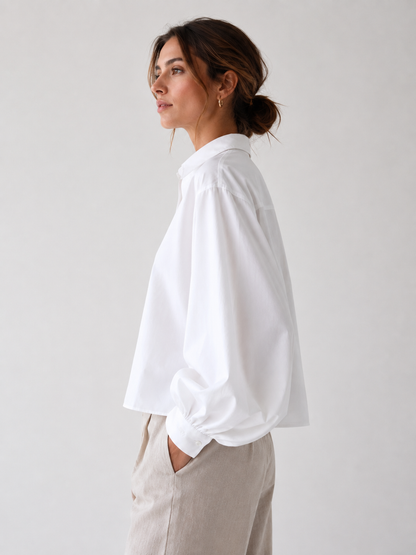 Eine Frau mit braunen Haaren, die zu einem tief sitzenden Dutt zusammengebunden sind, trägt eine locker sitzende weiße Bluse „Antonia Short White“ und eine hellbeige Hose. Sie steht im Profil vor einem schlichten, hellen Hintergrund, hat eine Hand in der Tasche und blickt nach vorne.