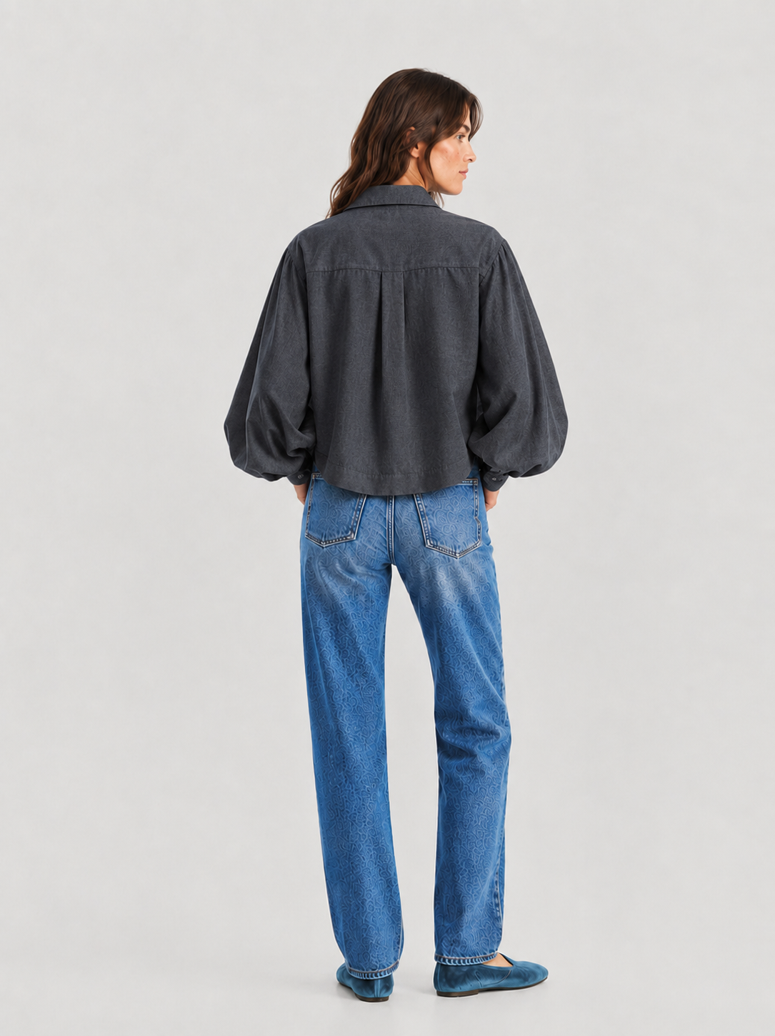 Eine Frau steht mit dem Rücken zur Kamera und trägt eine locker sitzende Bluse „Antonia Short Flanell Anthrazit“ mit langen Ärmeln, dazu eine blaue Jeans und türkisfarbene Ballerinas, vor einem schlichten, hellen Hintergrund.