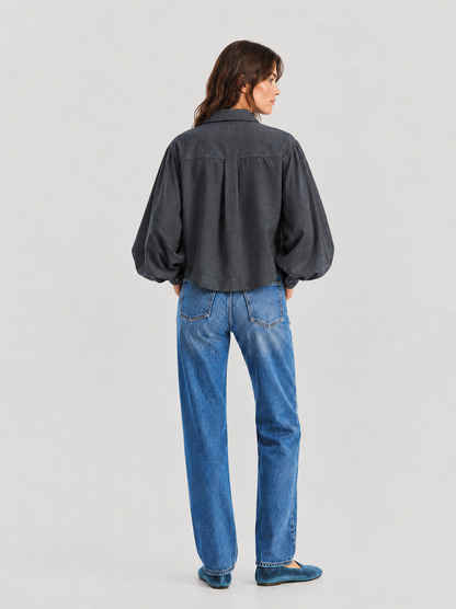 Eine Frau steht mit dem Rücken zur Kamera und trägt eine locker sitzende Bluse „Antonia Short Flanell Anthrazit“ mit langen Ärmeln, dazu eine blaue Jeans und türkisfarbene Ballerinas, vor einem schlichten, hellen Hintergrund.