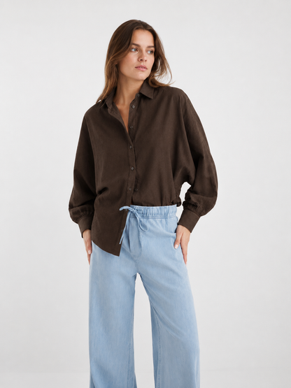 Eine Frau, die eine locker sitzende Bluse „Antonia Slim Espresso“ mit Ballonärmeln trägt, die in eine hellblaue Hose mit Kordelzug gesteckt ist; sie steht vor einem schlichten hellen Hintergrund, die Hände in den Taschen, und blickt leicht zur Seite.