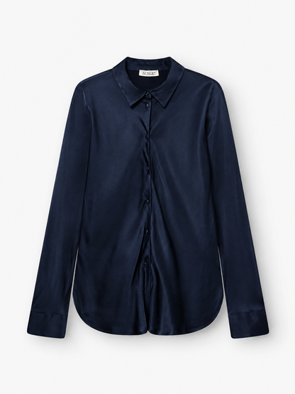 Die Bluse „Kaia Dunkelblau“ – ein langärmeliges, marineblaues Satinhemd mit Knopfleiste, spitzem Kragen und Manschetten – wird vor einem schlichten weißen Hintergrund präsentiert. Diese in Portugal gefertigte, elegante, fließende Bluse strahlt zeitlose Raffinesse und hochwertige Handwerkskunst aus.