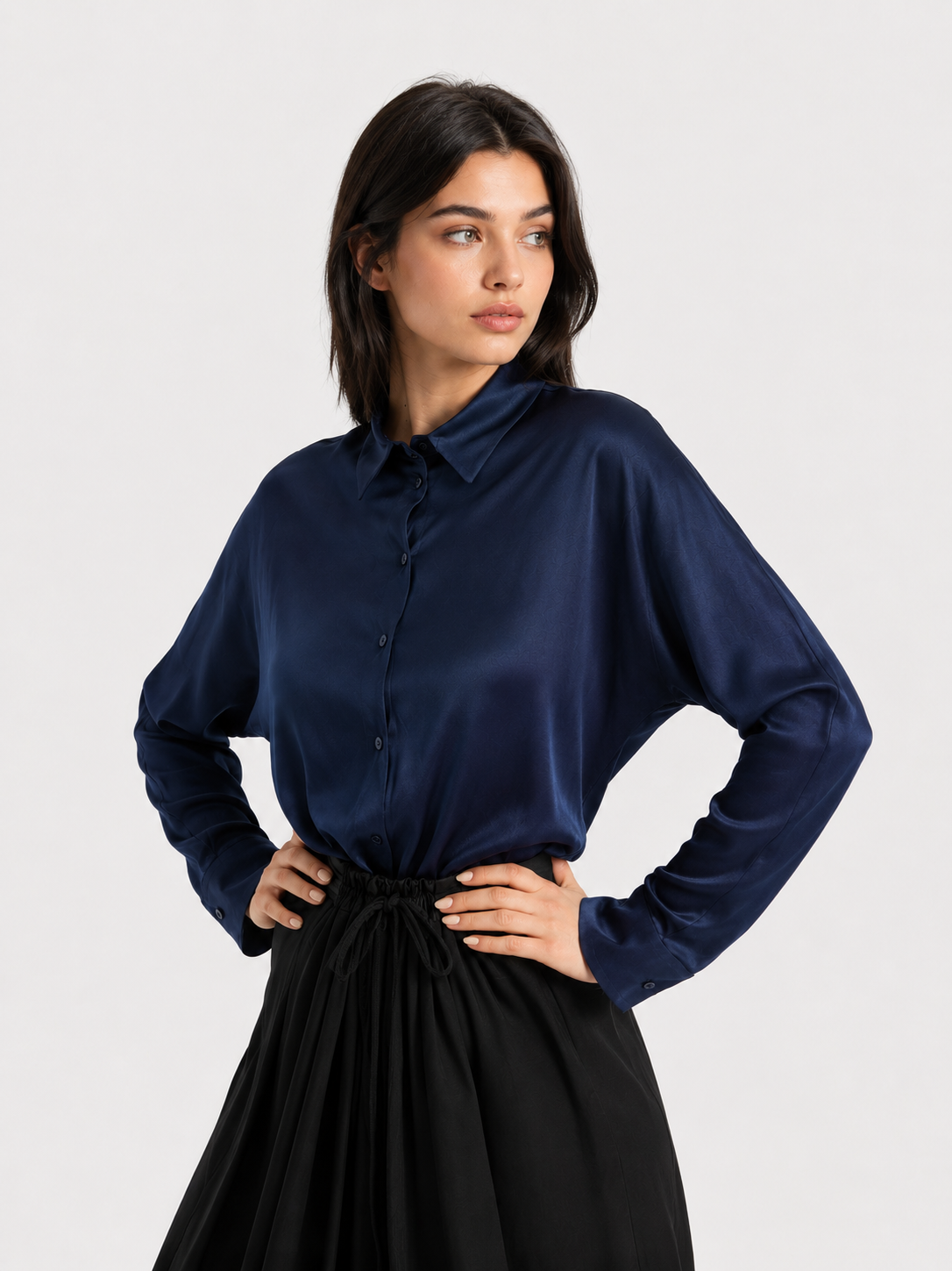 Eine Frau mit dunklem Haar trägt die Bluse „Kaia Dunkelblau“ – eine elegante, marineblaue Satinbluse mit Knöpfen –, dazu einen schwarzen Rock. Sie steht mit den Händen in den Hüften und blickt vor einem schlichten, hellen Hintergrund zur Seite. Diese fließend fallende Bluse wurde in Portugal hergestellt.