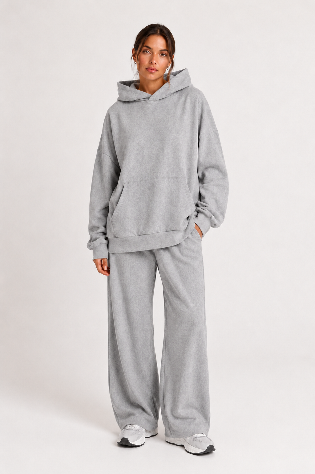 Vor einem schlichten Hintergrund steht eine Person, die den „Grauer Oversized Hoodie“ in Hellgrau trägt, dazu eine passende Sweathose mit weitem Bein und weiße Sneaker. Dieses in Portugal hergestellte, bequeme Outfit strahlt eine entspannte Atmosphäre aus, während die Person die Hände an den Seiten ruhen lässt und in die Kamera blickt.