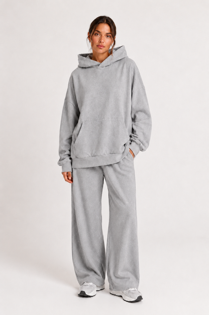 Eine Person steht vor einem schlichten Hintergrund und trägt ein hellgraues, übergroßes Jogger-Kapuzensweatshirt sowie eine dazu passende Jogger-Hose mit weitem Bein, dazu weiße Sneaker.
