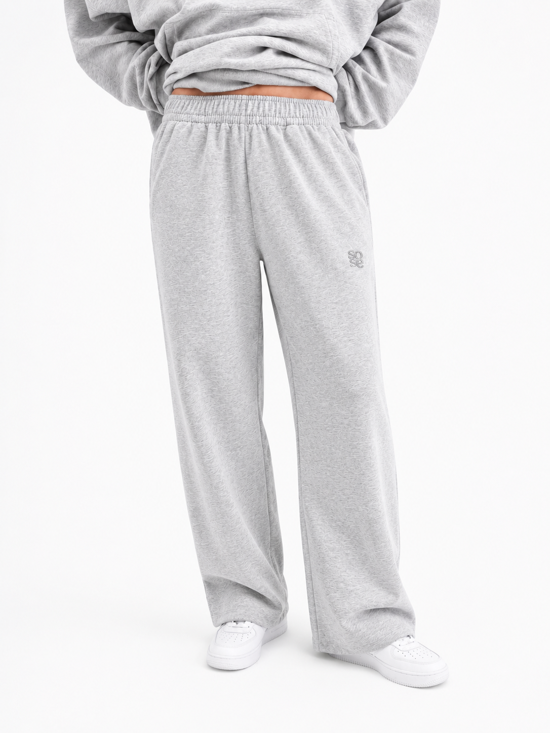 Eine Person ist von der Taille abwärts zu sehen, die eine hellgraue Jogginghose trägt, dazu ein passendes Sweatshirt und weiße Sneaker, und vor einem schlichten weißen Hintergrund steht.
