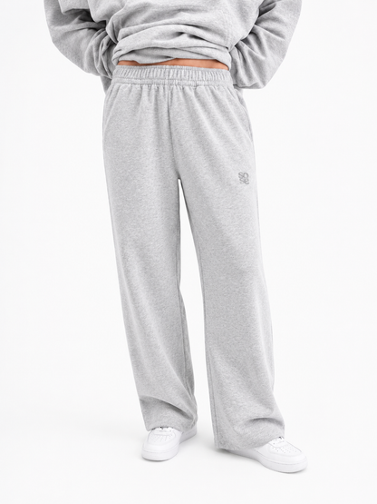 Eine Person ist von der Taille abwärts zu sehen, die eine hellgraue Jogginghose trägt, dazu ein passendes Sweatshirt und weiße Sneaker, und vor einem schlichten weißen Hintergrund steht.