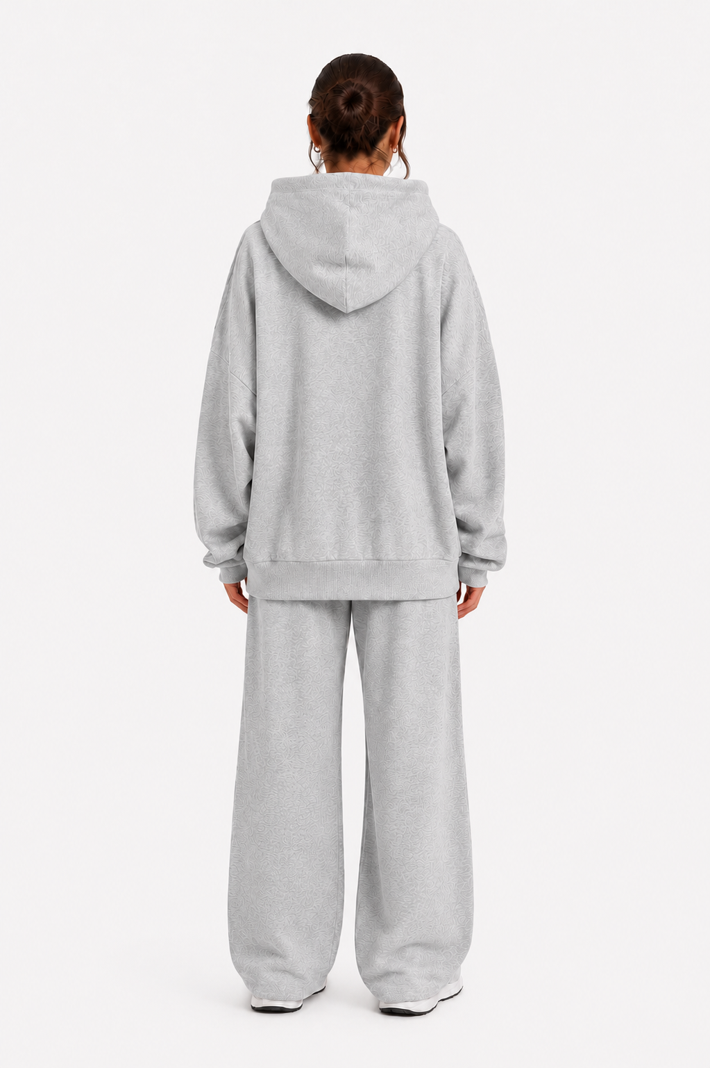 Eine Person steht mit dem Rücken zur Kamera vor einem schlichten weißen Hintergrund und trägt das „Jogger“-Set – ein übergroßes, hellgraues Sweatshirt mit Kapuze und eine dazu passende Jogginghose, die aus weichem Fleece in Portugal gefertigt wurden.