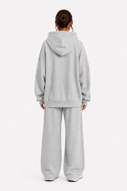 Eine Person steht mit dem Rücken zur Kamera vor einem schlichten weißen Hintergrund und trägt das „Jogger“-Set – ein übergroßes, hellgraues Sweatshirt mit Kapuze und eine dazu passende Jogginghose, die aus weichem Fleece in Portugal gefertigt wurden.