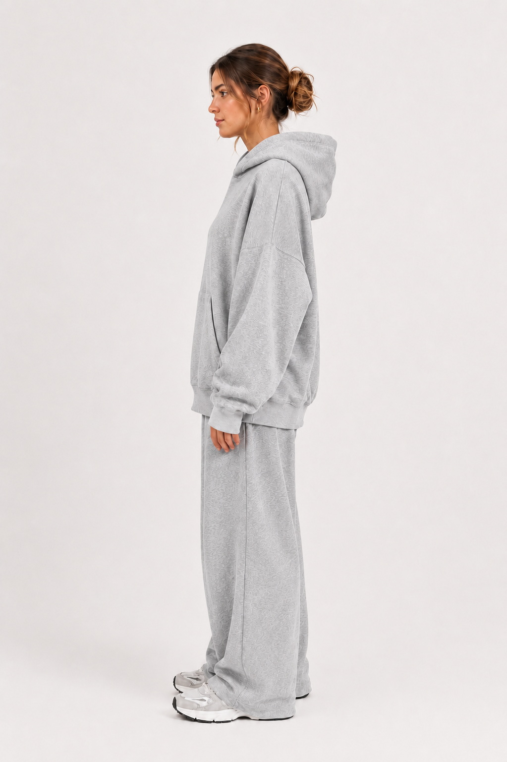Vor einem schlichten weißen Hintergrund steht eine Frau im Profil. Sie trägt einen übergroßen grauen Hoodie, dazu die „Jogger“ – eine in Portugal hergestellte Fleece-Jogginghose mit weitem Bein – sowie silberne Sneaker. Ihr Haar ist zu einem lockeren Dutt zusammengebunden, was den gemütlichen Look abrundet.