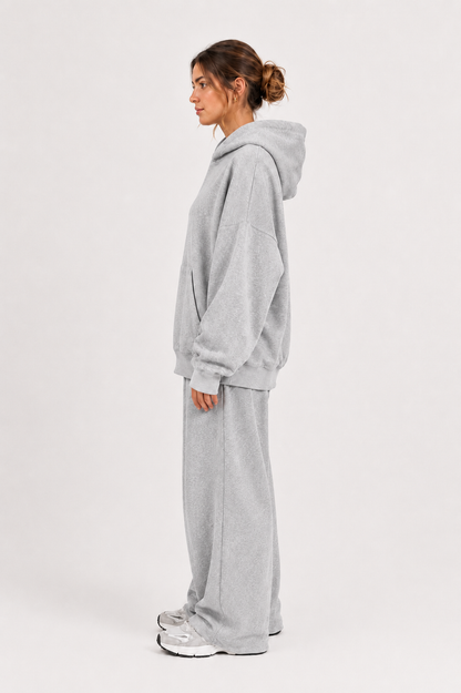 Vor einem schlichten weißen Hintergrund steht eine Frau im Profil. Sie trägt einen übergroßen grauen Hoodie, dazu die „Jogger“ – eine in Portugal hergestellte Fleece-Jogginghose mit weitem Bein – sowie silberne Sneaker. Ihr Haar ist zu einem lockeren Dutt zusammengebunden, was den gemütlichen Look abrundet.