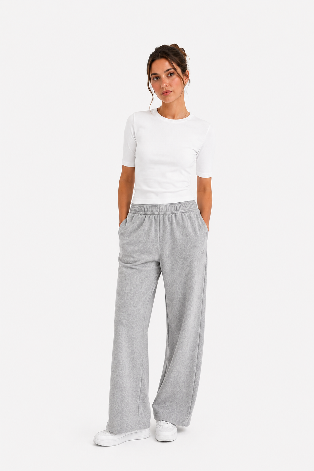 Eine Frau steht vor einem weißen Hintergrund und trägt ein eng anliegendes weißes T-Shirt mit kurzen Ärmeln, eine lockere graue Jogginghose mit weitem Bein aus recyceltem Polyester sowie weiße Sneaker; sie hat die Hände in den Taschen und einen neutralen Gesichtsausdruck.