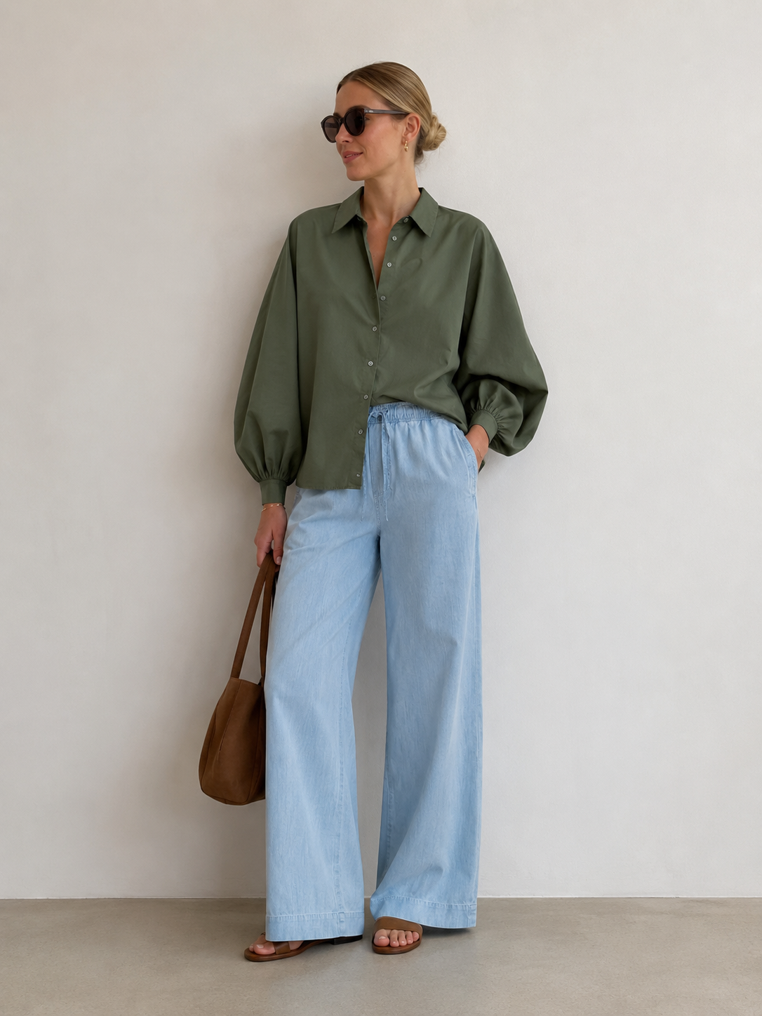 Eine Frau steht vor einer schlichten Wand und trägt eine Sonnenbrille, die olivgrüne Bluse „Antonia Olive“ aus Baumwolle mit Ballonärmeln (Einheitsgröße), eine hellblaue Hose mit weitem Bein, braune Sandalen und hält eine braune Umhängetasche in der Hand.