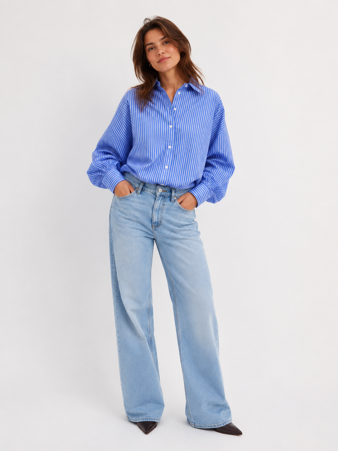 Eine Frau mit dunkelbraunen Haaren steht mit den Händen in den Taschen da. Sie trägt die „Bluse Antonia Slim Ocean“ – eine blau-weiß gestreifte Bluse in schmaler Passform –, dazu eine hellblaue Jeans mit weitem Bein und schwarze spitze Schuhe. Der Hintergrund ist schlicht weiß.