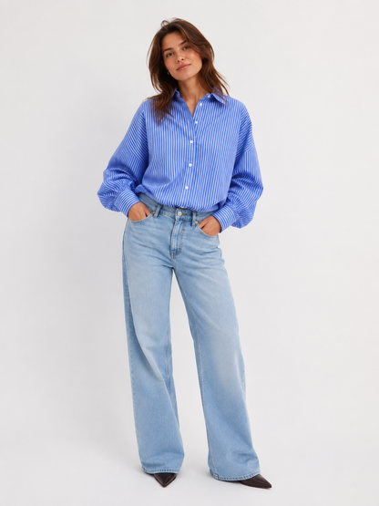 Eine Frau mit dunkelbraunen Haaren steht mit den Händen in den Taschen da. Sie trägt die „Bluse Antonia Slim Ocean“ – eine blau-weiß gestreifte Bluse in schmaler Passform –, dazu eine hellblaue Jeans mit weitem Bein und schwarze spitze Schuhe. Der Hintergrund ist schlicht weiß.