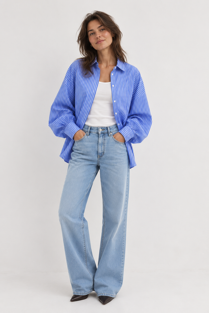 Eine Frau steht vor einem weißen Hintergrund und trägt die Bluse „Antonia Slim Ocean“, ein blau gestreiftes Hemd mit Knöpfen über einem weißen Oberteil, dazu hellblaue Jeans mit weitem Bein und schwarze spitze Schuhe. Ihre Hände stecken in den Taschen, und ihr Gesichtsausdruck wirkt entspannt.