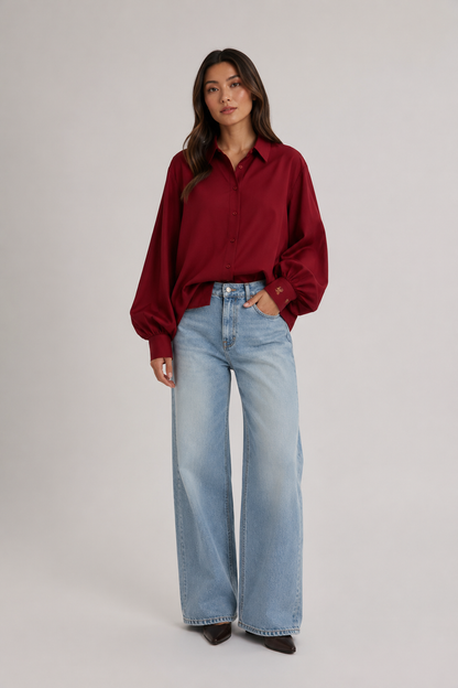 Vor einem schlichten Hintergrund steht eine Frau, die eine locker sitzende Bluse „Antonia Burgundy“ mit Ballonärmeln trägt, dazu hellblaue Jeans mit weitem Bein und spitze braune Schuhe.